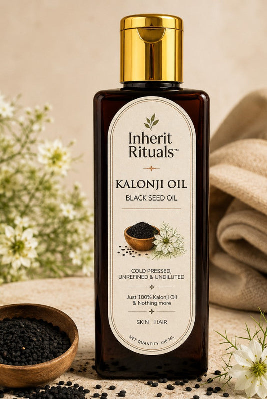 A bottle of Inherit Rituals Kalonji Oil (Black Seed Oil) with a gold cap and cream-colored label, set on a beige fabric surface with black seeds, a small wooden bowl, and white flowers.